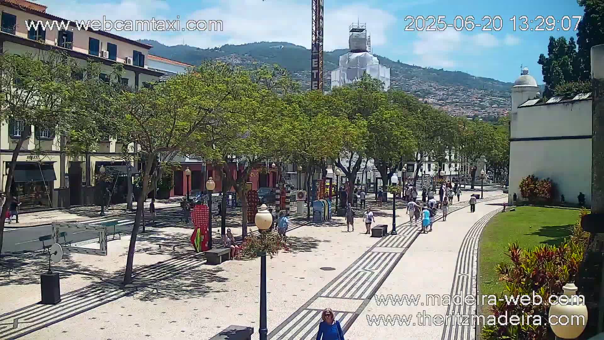 A sunny city street scene shows pedestrians strolling along a patterned walkway lined with trees and buildings, with a mountain visible in the background.