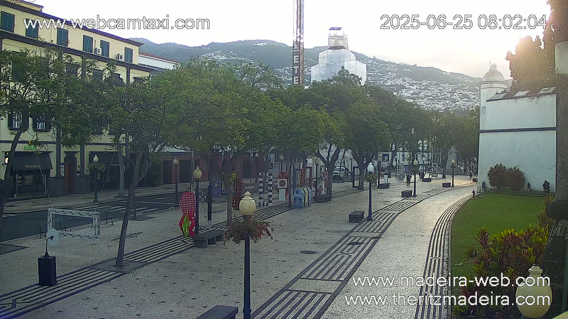 A sunlit plaza with a patterned walkway, lined with trees and buildings, in front of a hill with houses on it.