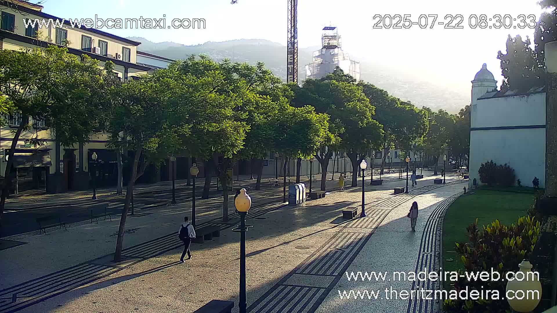A sunlit paved walkway lined with trees and buildings, with a few pedestrians visible.