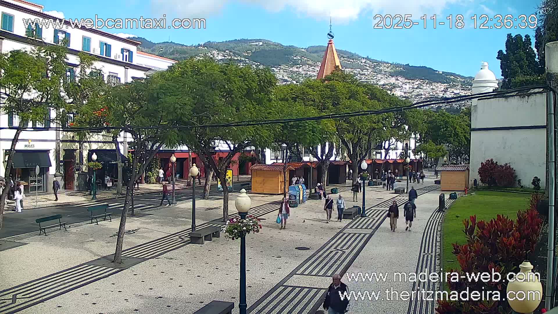 A sunlit paved walkway lined with trees and buildings, with a few pedestrians visible.