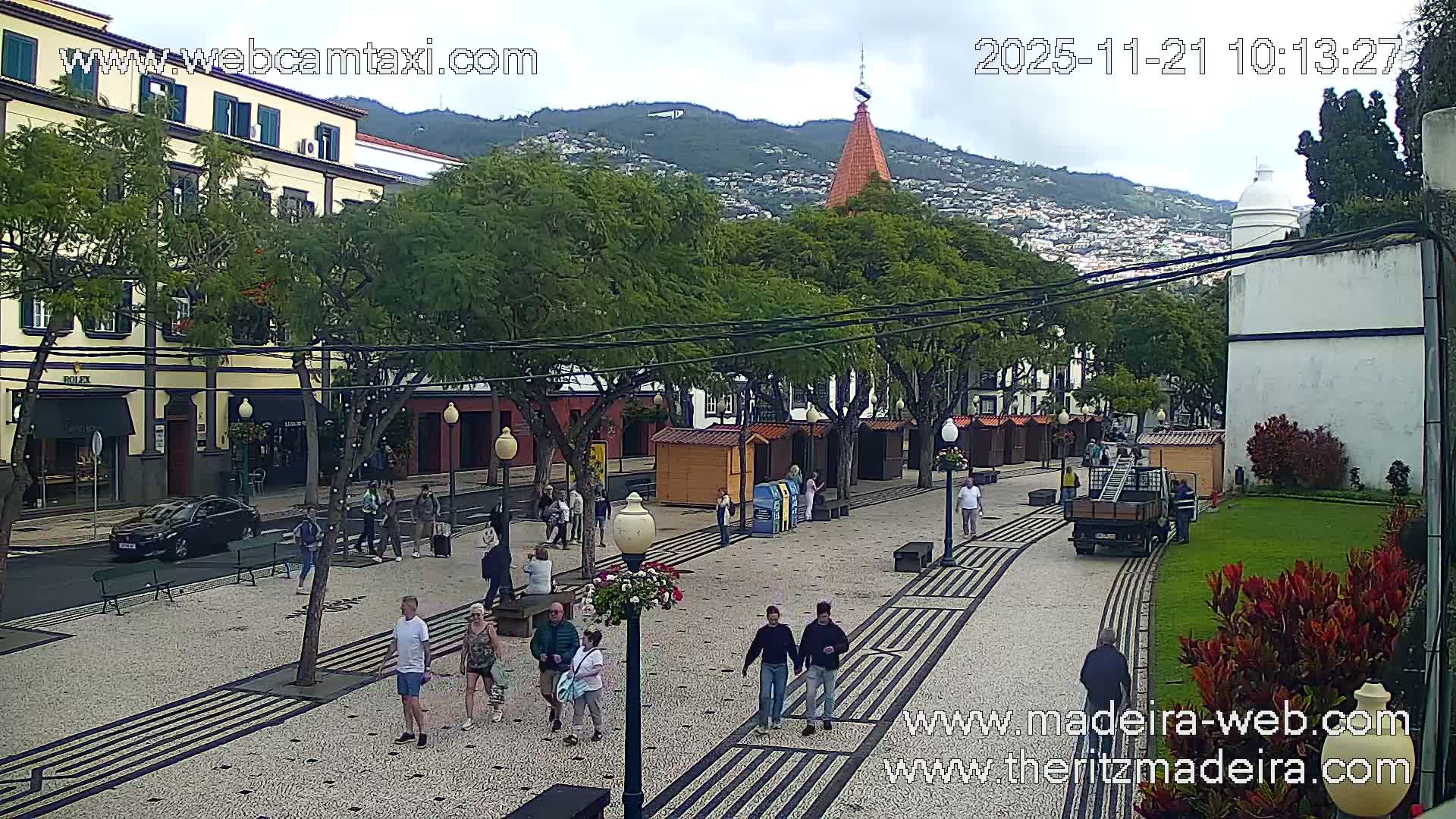 On an overcast day, a wide pedestrian street featuring patterned paving, numerous people walking, and small market stalls is visible, flanked by traditional buildings and trees, with a prominent orange conical-roofed tower and a hillside town in the background.