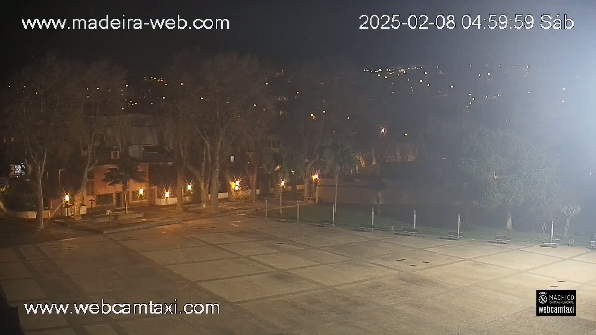 A nighttime view of an empty plaza, surrounded by trees and buildings with lights, under hazy conditions.