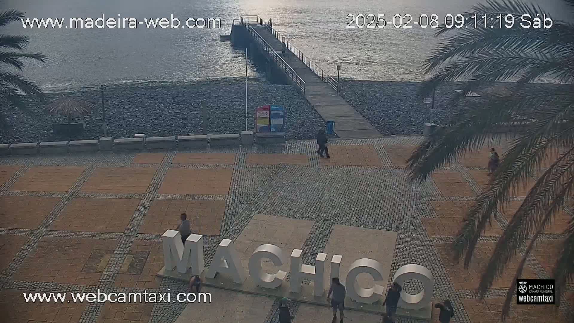 A paved plaza with a large "Machico" sign overlooks a pebble beach and a pier extending into a calm, gray ocean under an overcast sky.