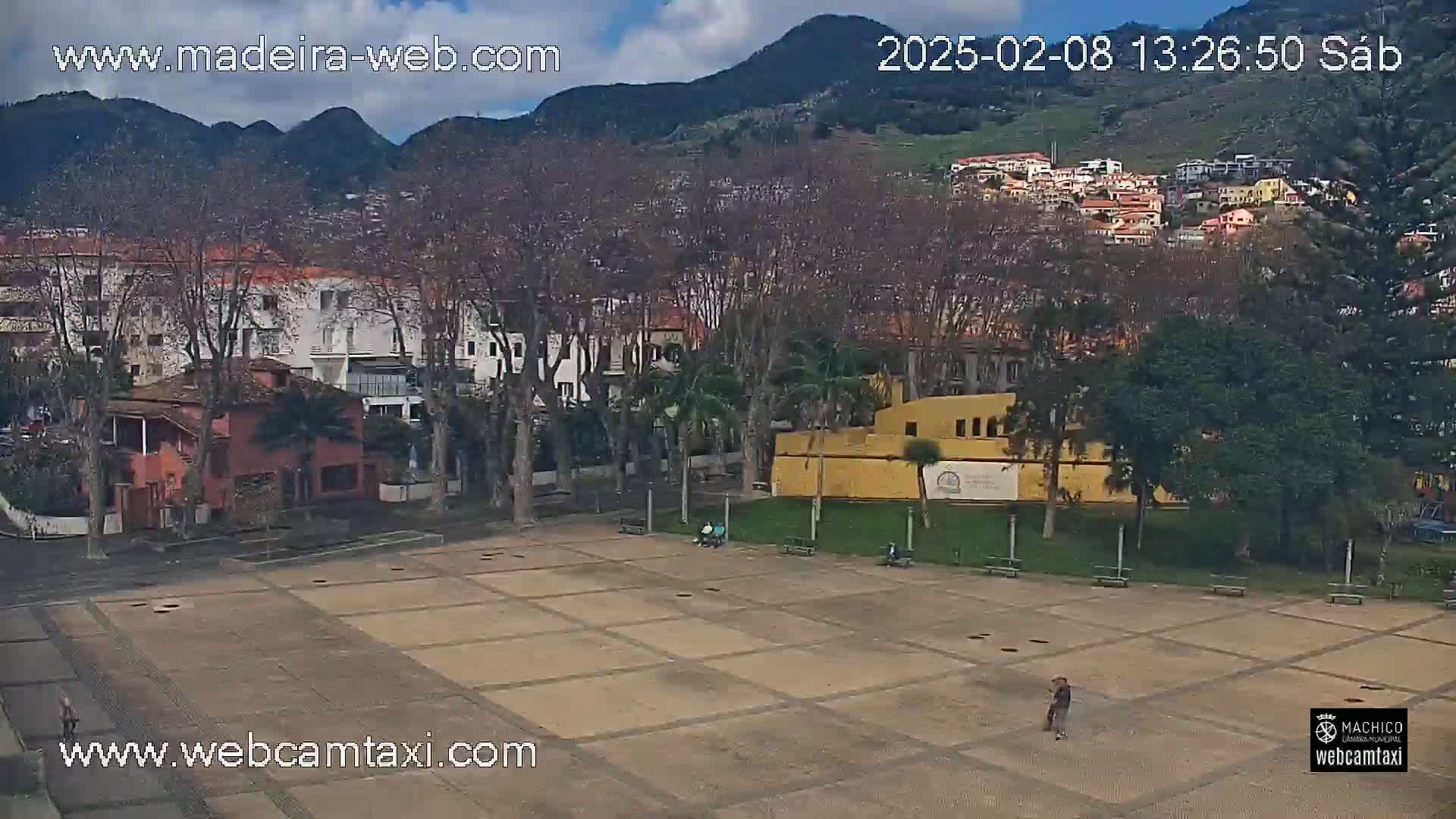 A mostly empty paved plaza sits before a town nestled against a mountainside under a partly cloudy sky.