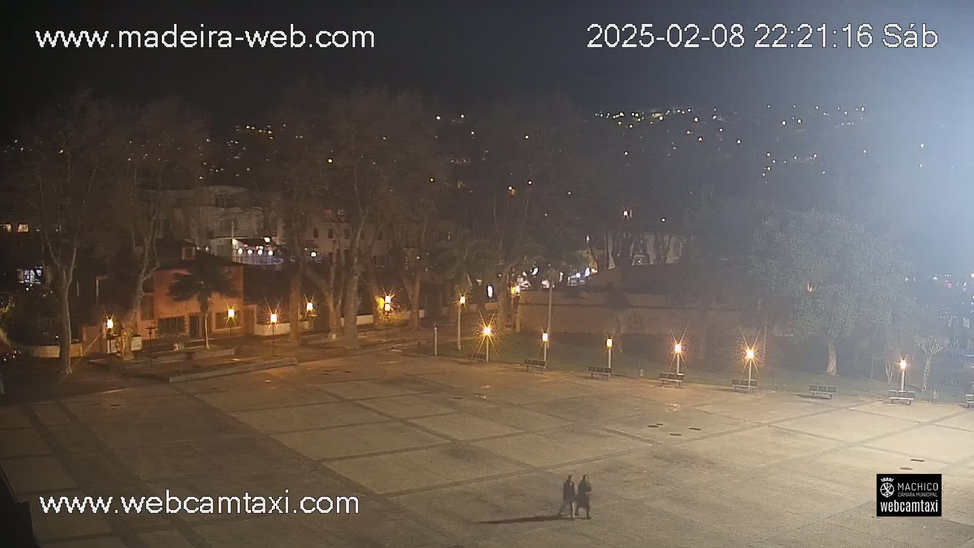 A nighttime view of an empty plaza, illuminated by lampposts, with trees and buildings in the background under a hazy sky.