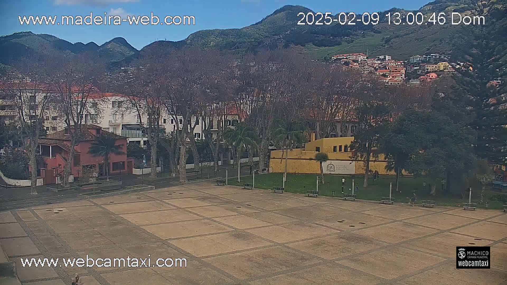 A paved plaza is surrounded by buildings, trees, and hills under a clear, sunny sky.