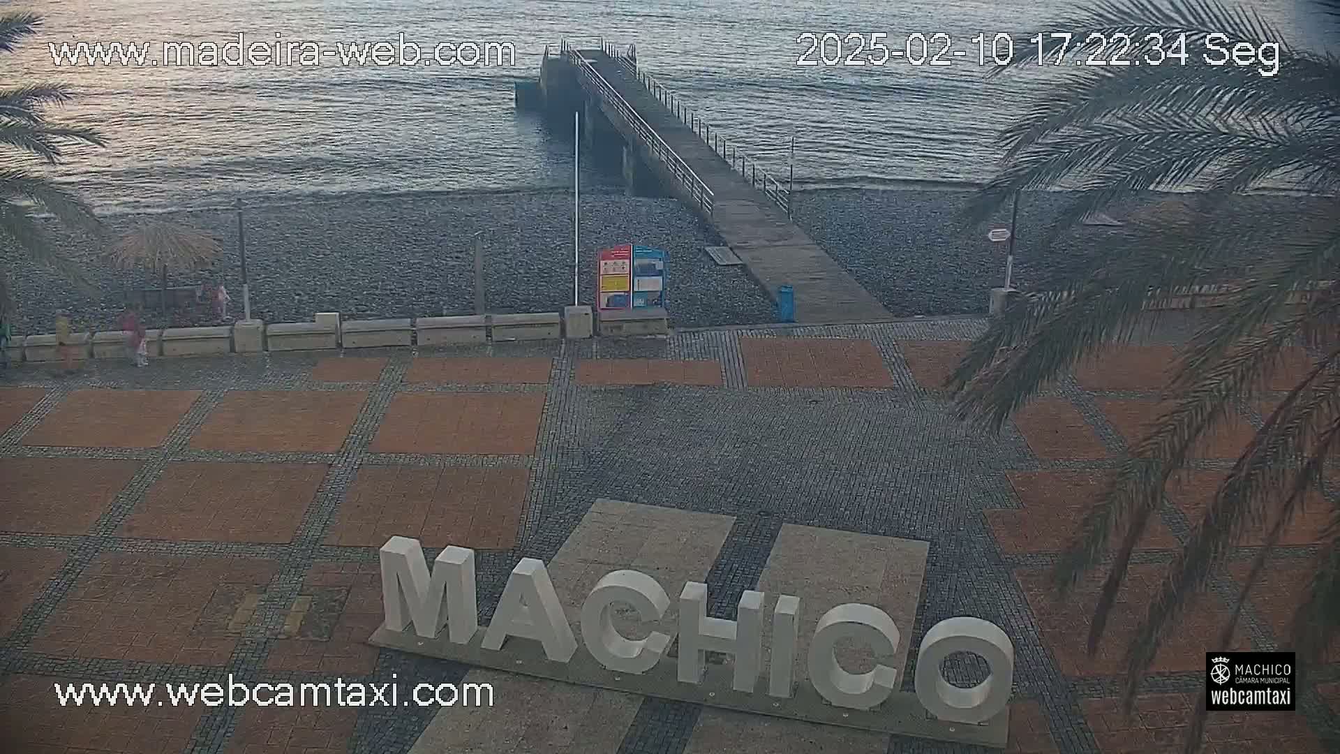 A paved area with the word "Machico" in large letters overlooks a pebble beach and a pier extending into calm water under a partly cloudy sky.
