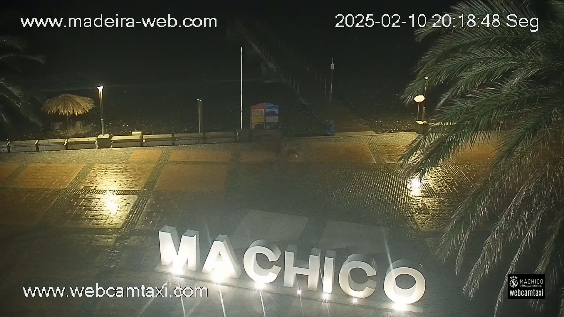 A nighttime view of a wet paved area with the illuminated word "MACHICO" in large letters, palm trees, and a beach in the background.