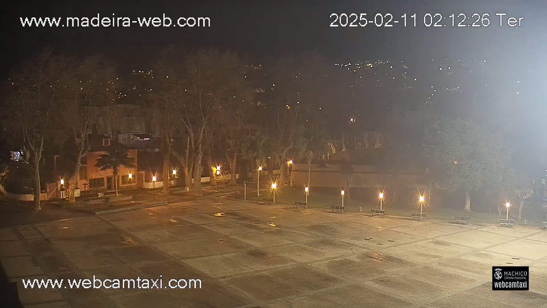 A nighttime view of a mostly empty plaza, surrounded by trees and buildings under a hazy sky.
