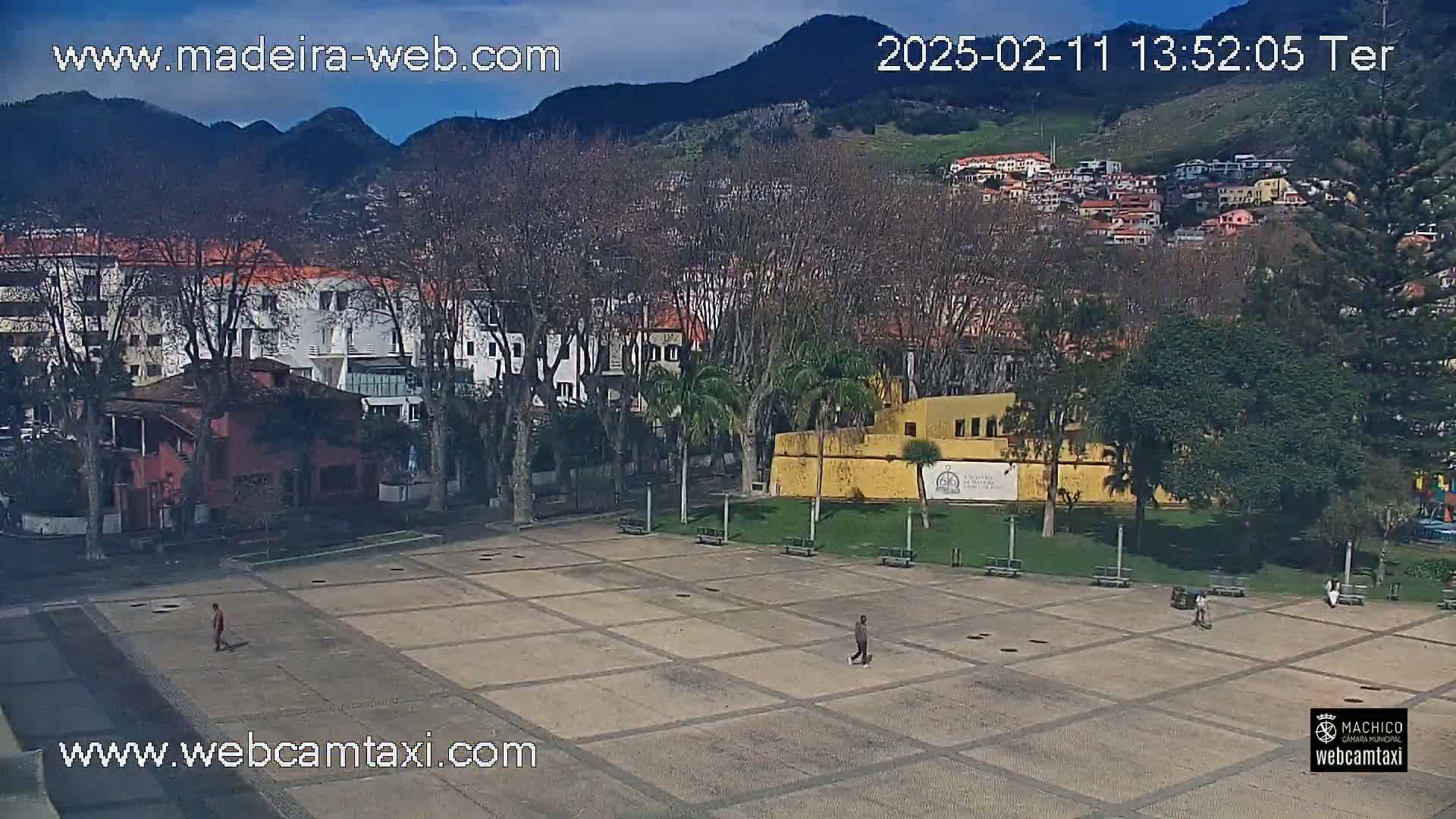 A paved plaza is surrounded by buildings and trees, with mountains in the background under a mostly clear sky.