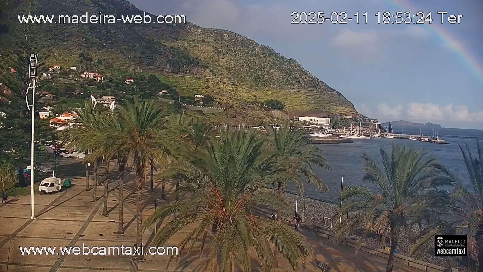 A harbor filled with boats sits below a hillside town nestled against a steep, green mountain, all under a partly cloudy sky with a rainbow visible.