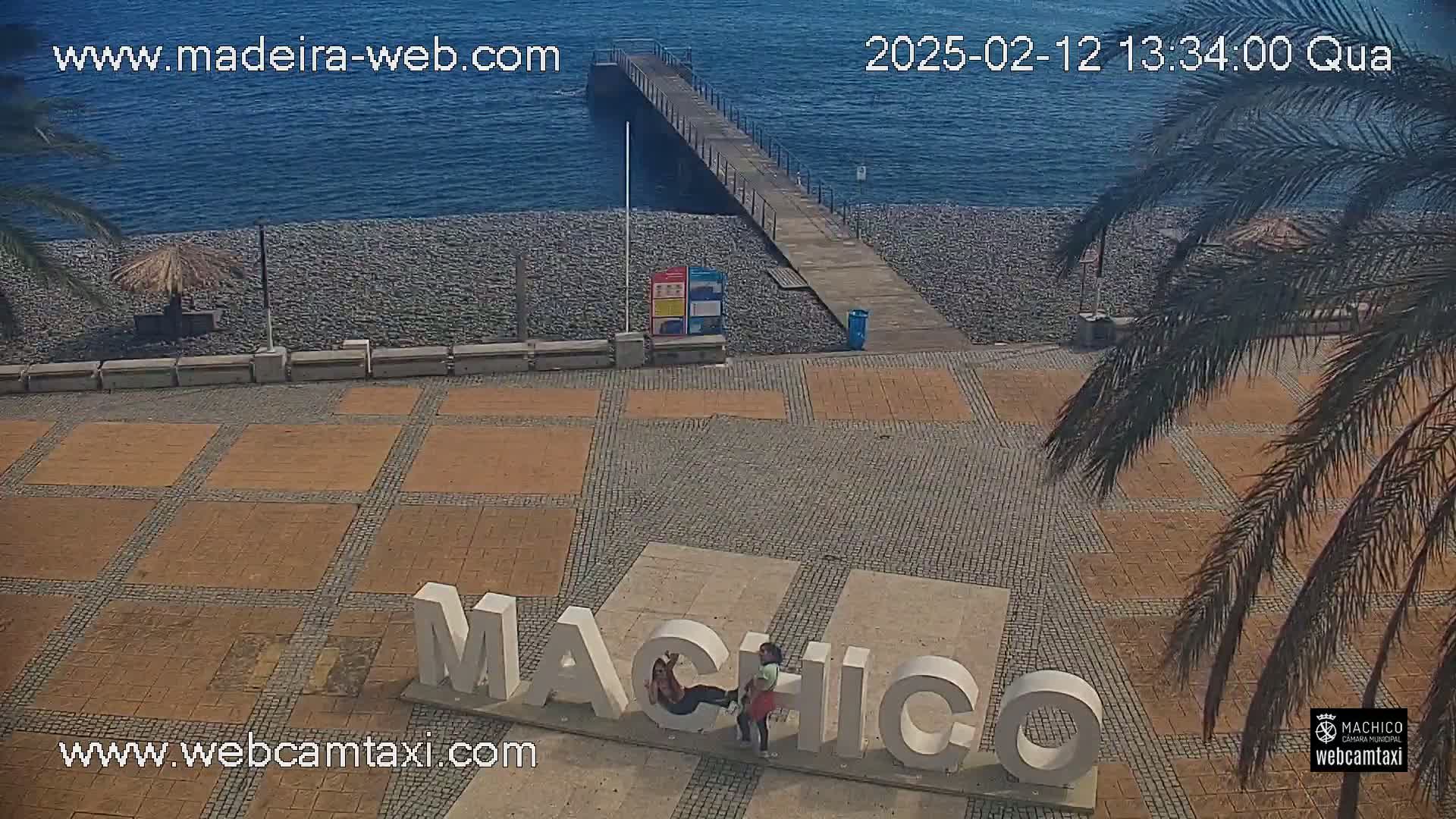 A beachside plaza with a pebble beach, a pier, palm trees, and large letters spelling out a place name, under a sunny sky.