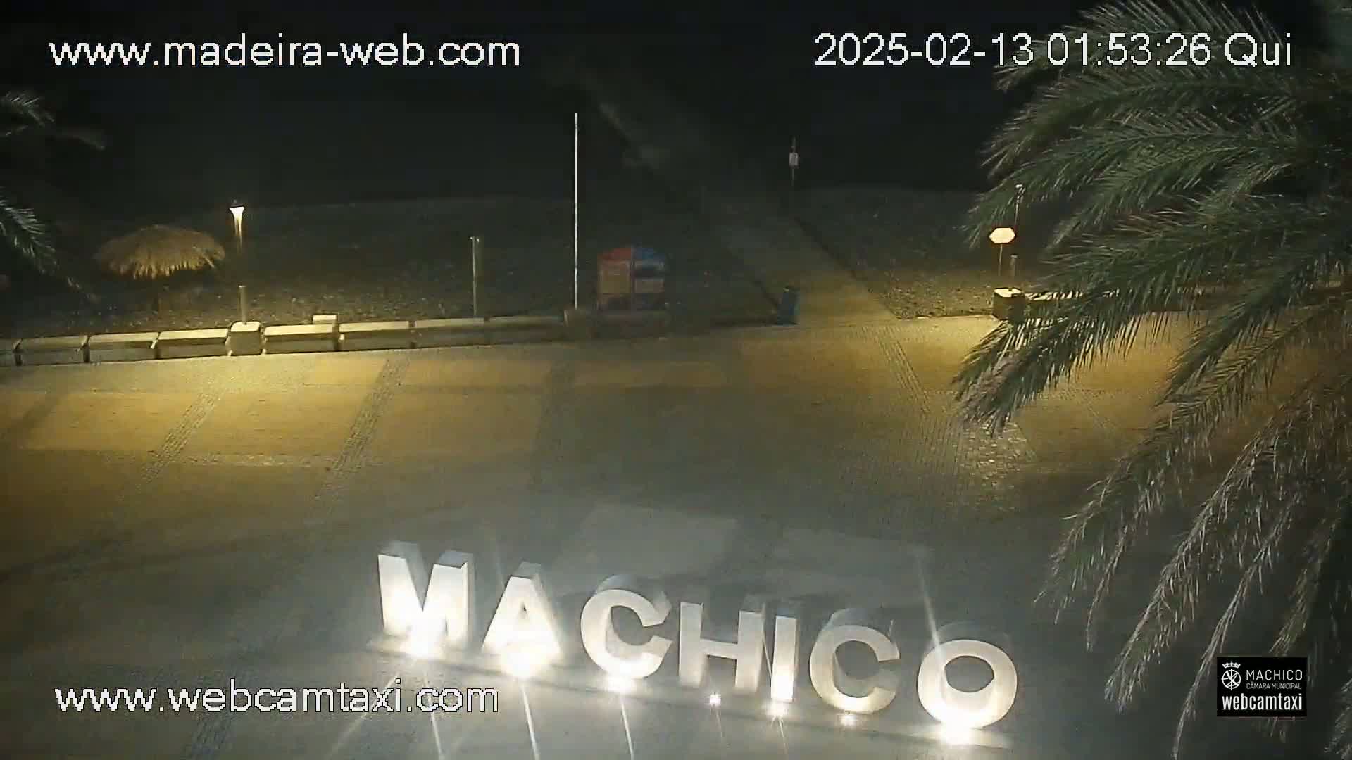 A nighttime view of a paved area with palm trees, a large illuminated sign spelling "Machico," and a small thatched umbrella.