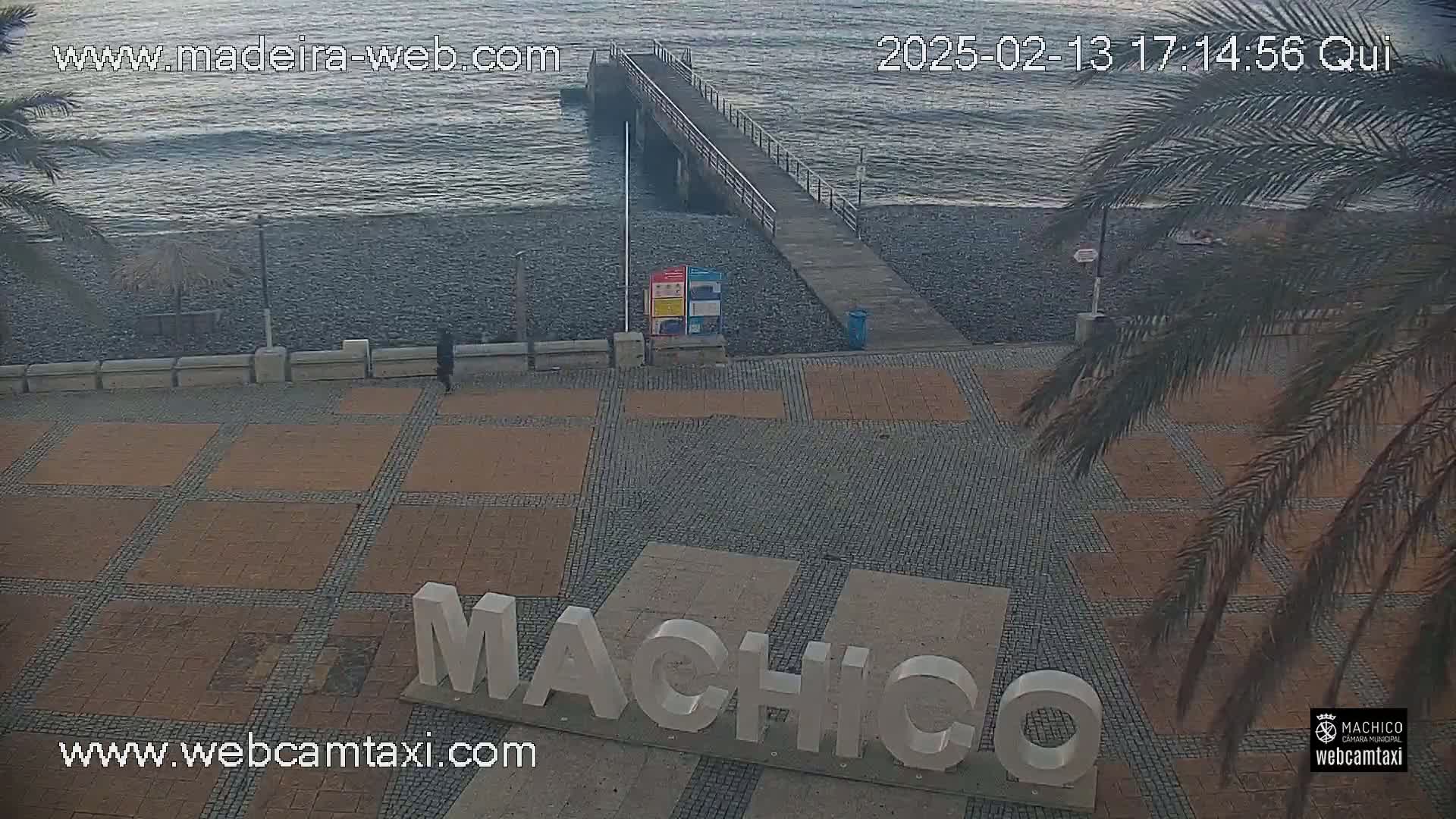 A person walks along a paved plaza near a pebble beach and pier on an overcast day.