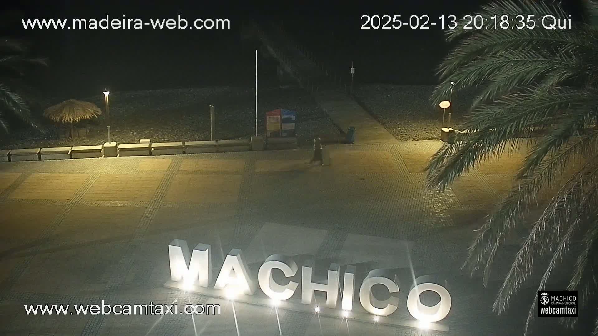 A nighttime view of a paved area with the word "MACHICO" displayed in large letters, near a beach with palm trees under a clear sky.