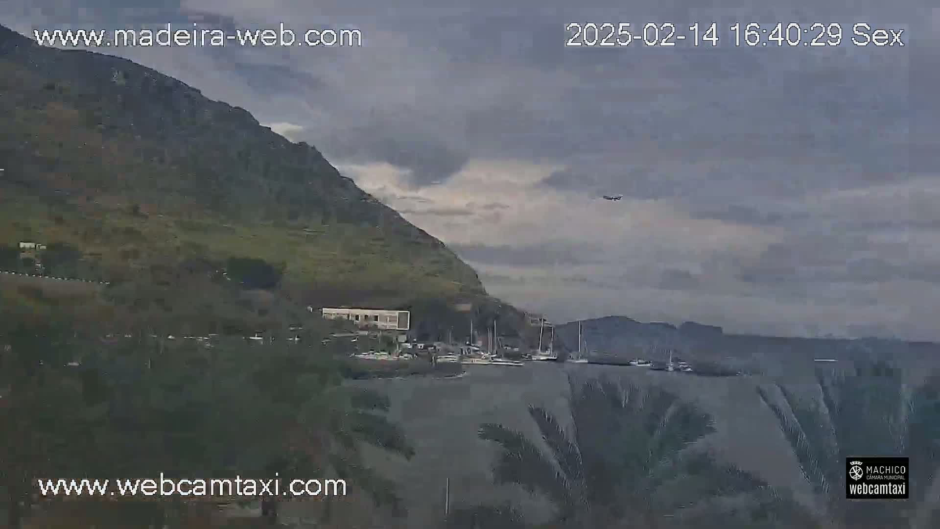 A plane flies over a harbor nestled beneath a large, green hill under a cloudy sky.