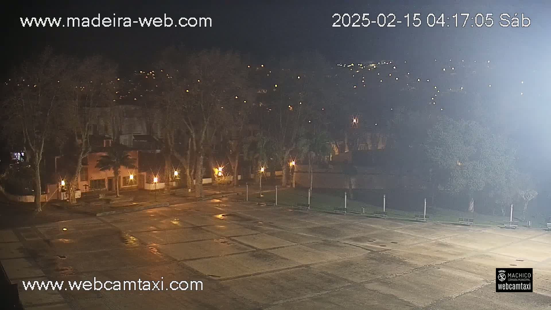 A nighttime view of a mostly empty plaza, bordered by trees and buildings, under a hazy sky.