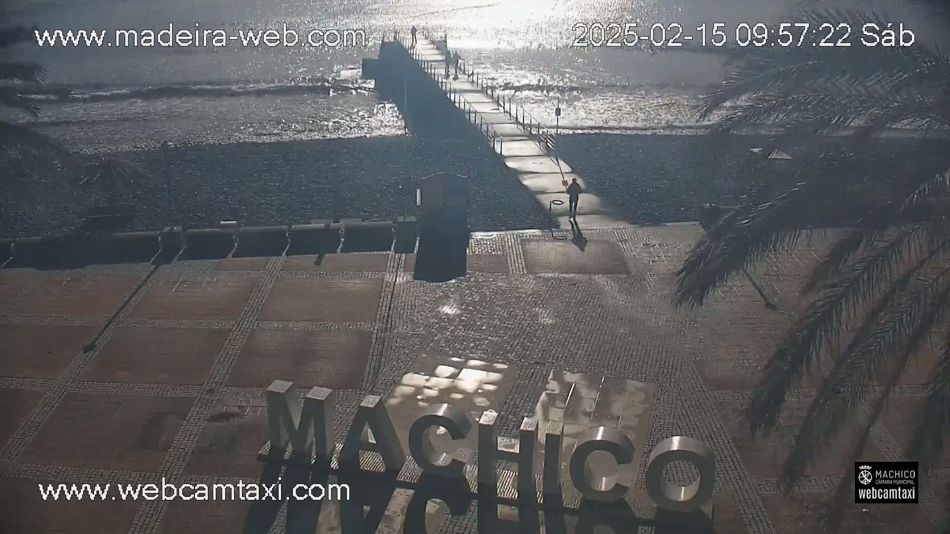 A paved area with a large "Machico" sign near a pebble beach and a pier extending into a calm, sunlit sea.