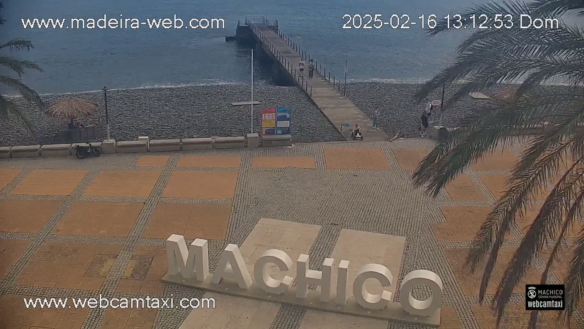 A seaside plaza with a pier, a pebble beach, and large white letters spelling out a place name, under a mostly clear sky.