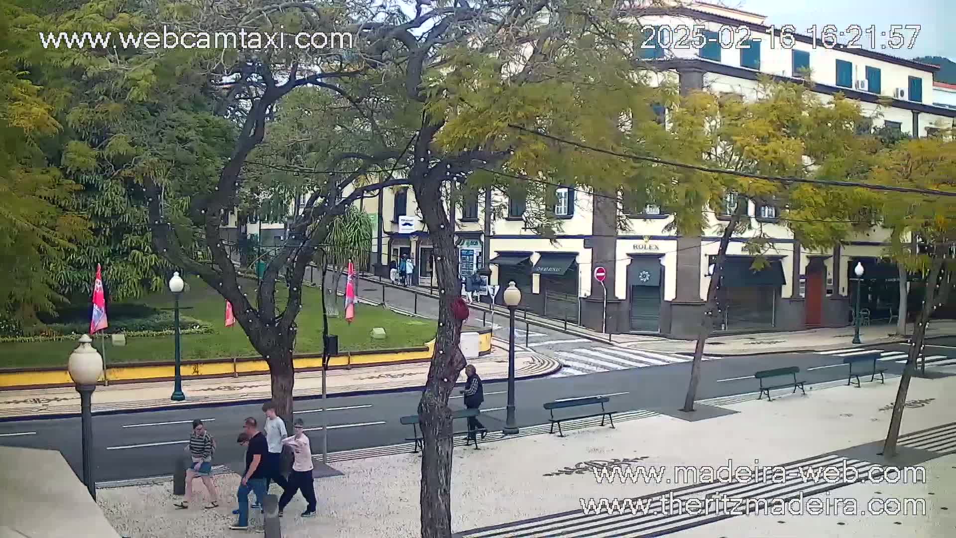 A town square with several people walking, benches, and a building with shops, under an overcast sky.