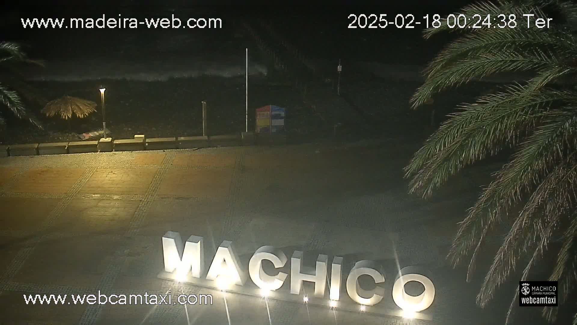 A nighttime view of a paved area with a large illuminated sign that reads "MACHICO," near a dark ocean with waves, under partly cloudy skies.