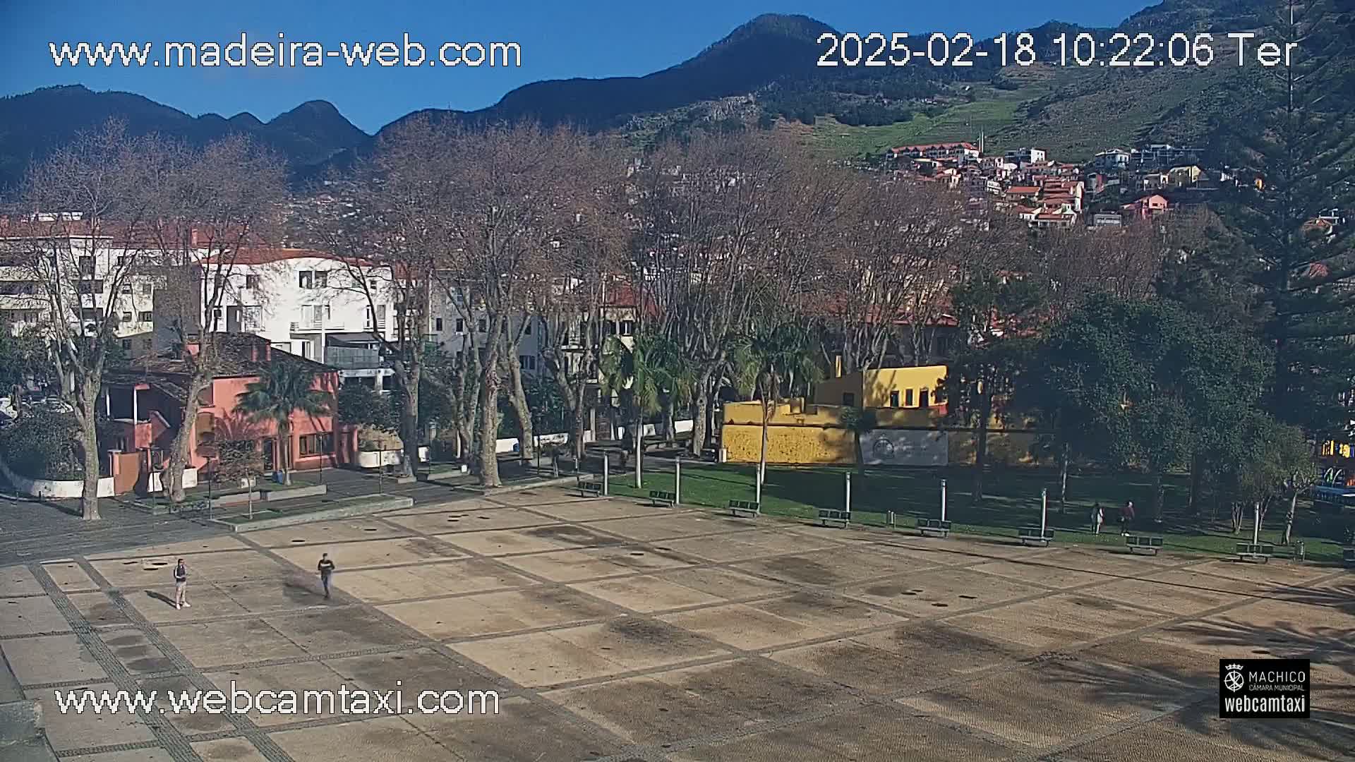 A large, empty plaza with a tiled surface is surrounded by buildings, trees, and a small grassy area, all under a clear, sunny sky with mountains in the background.
