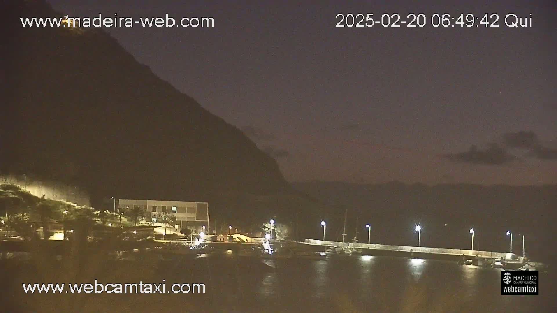 A nighttime view of a calm body of water with a small building and docked boats near a dark mountain under a mostly clear, dusky sky.