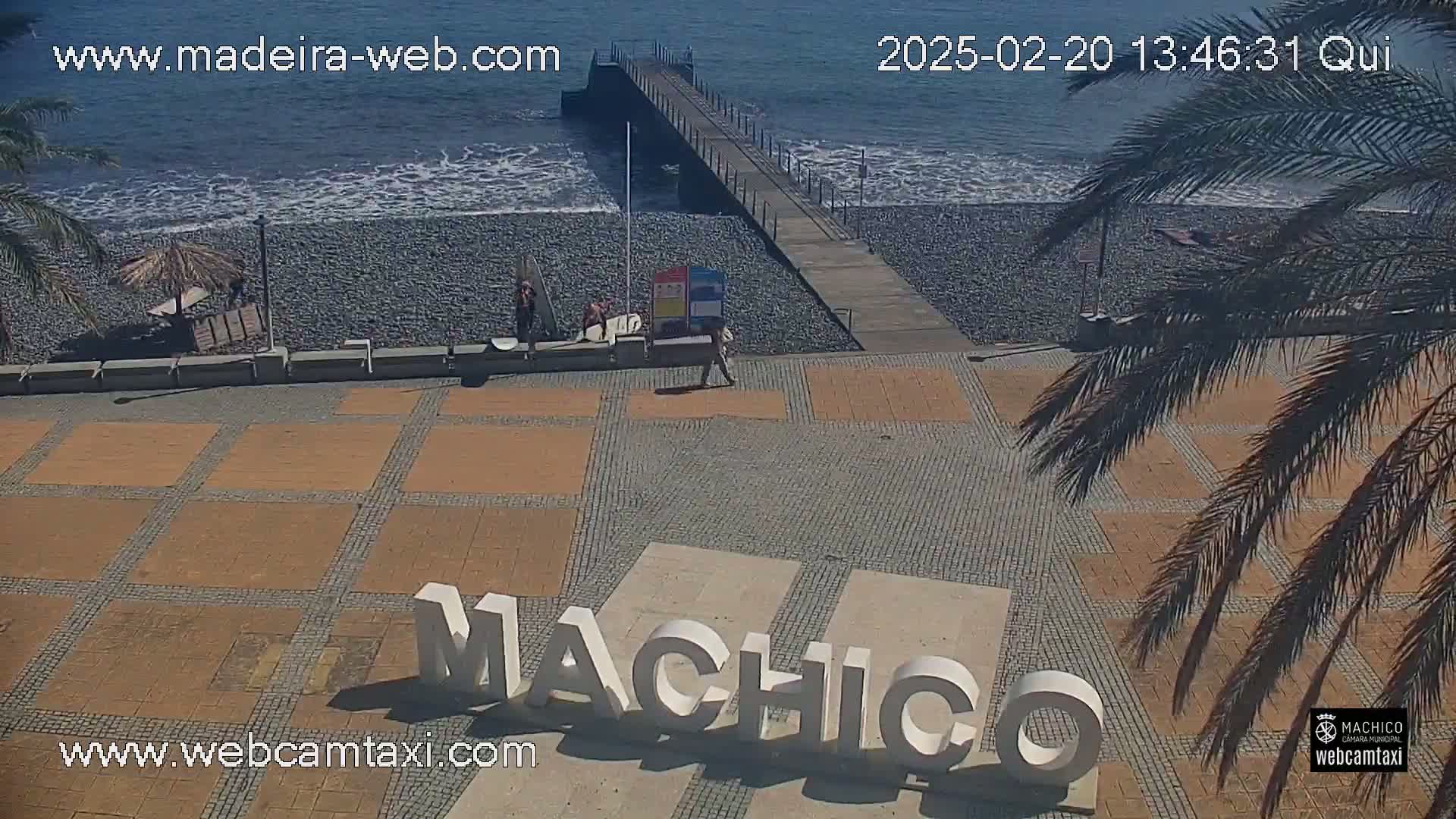 A beach with a pebble shoreline and a long pier, under sunny skies, features a paved plaza with large lettering spelling out "Machico."
