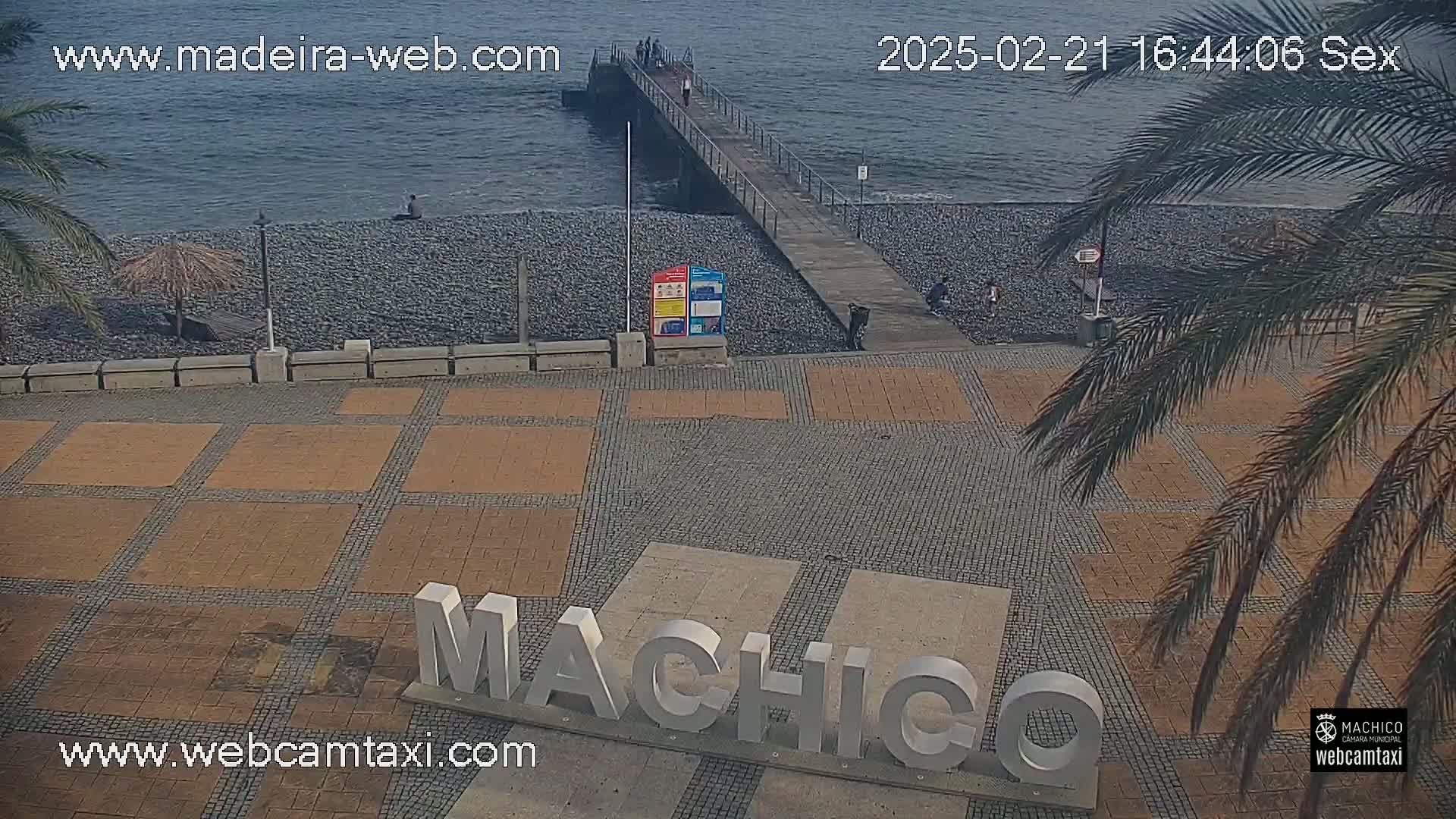 A paved plaza with a large "Machico" sign sits next to a pebble beach and a pier extending into a calm ocean under a partly cloudy sky.