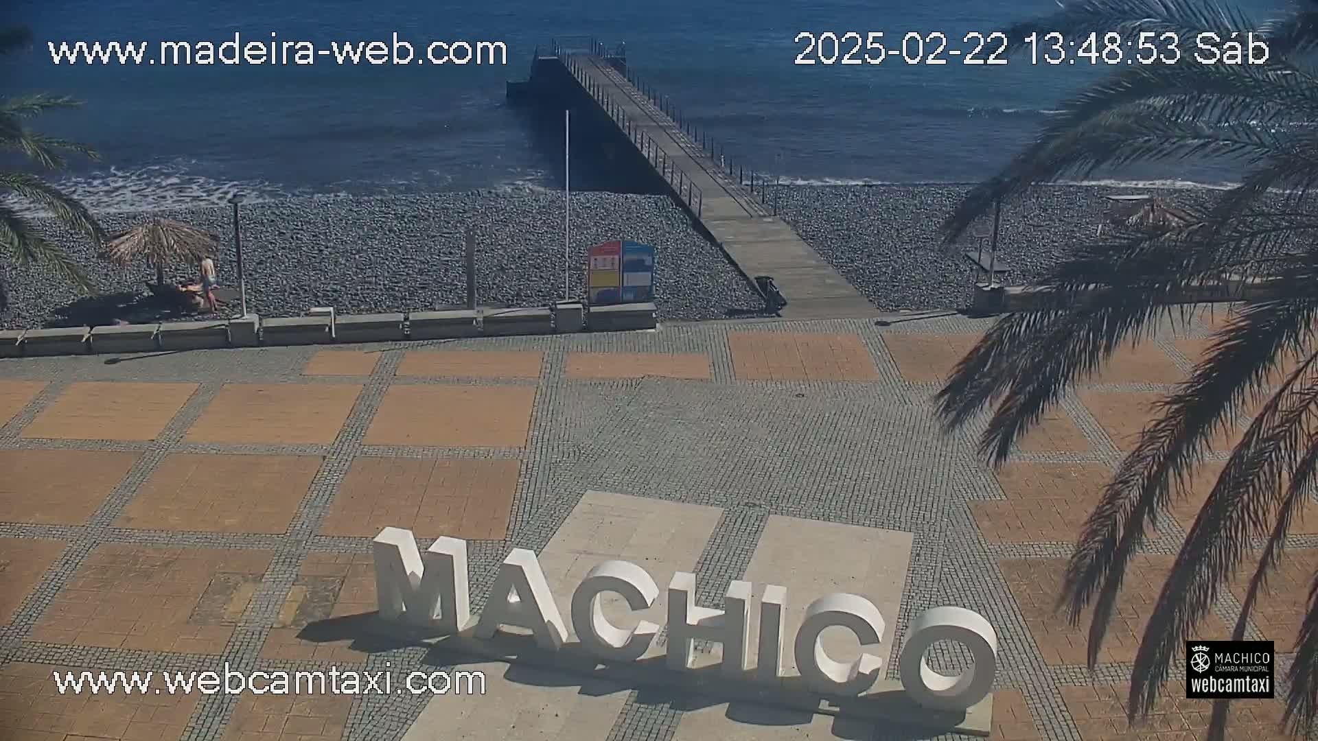 A sunny seaside plaza with a pebble beach, a pier, and large letters spelling out a place name.
