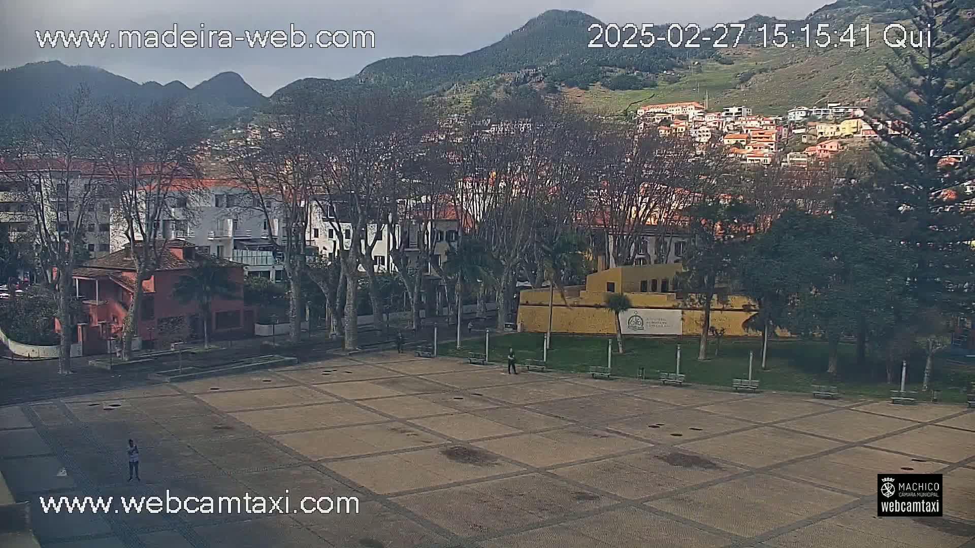 A mostly empty paved plaza is seen, with a few people, surrounded by buildings and trees under an overcast sky, mountains visible in the background.