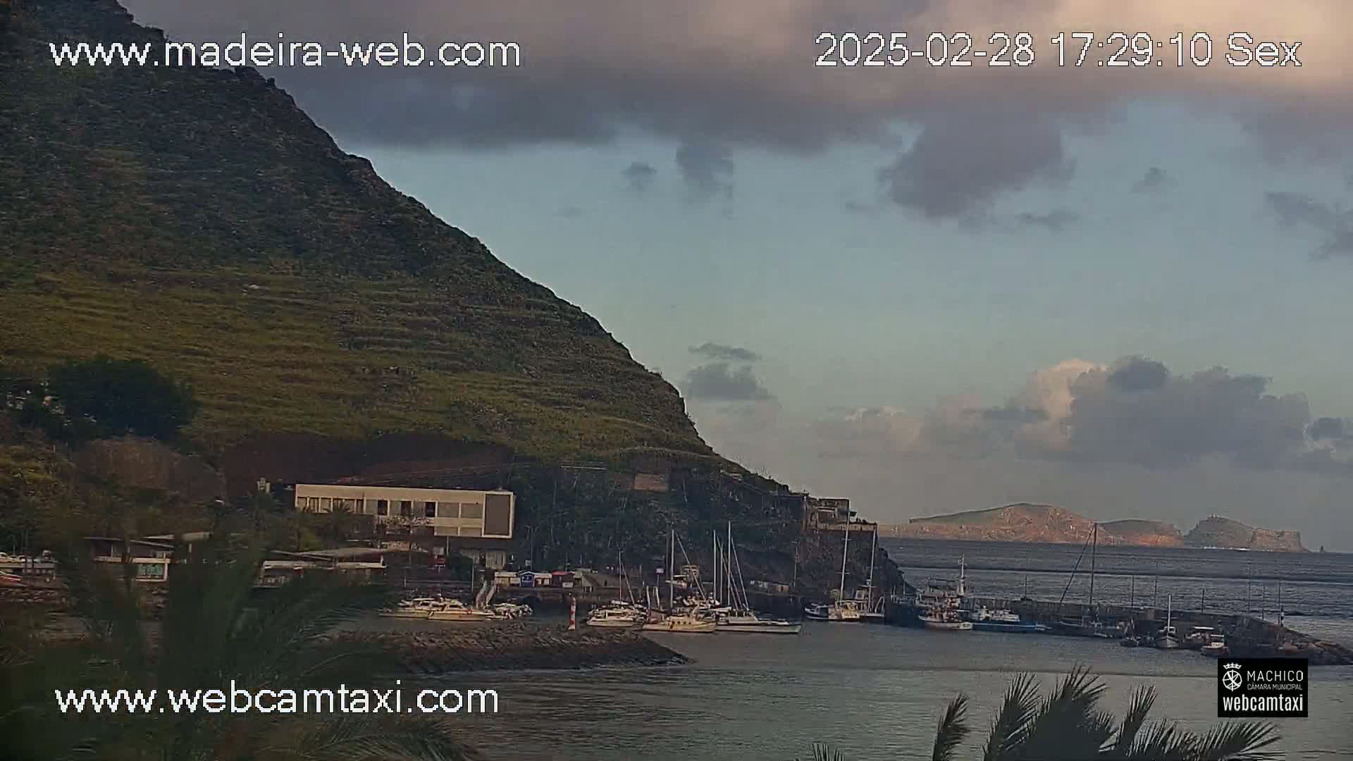 A partly cloudy sky overlooks a harbor filled with sailboats nestled beside a steep, terraced hillside.