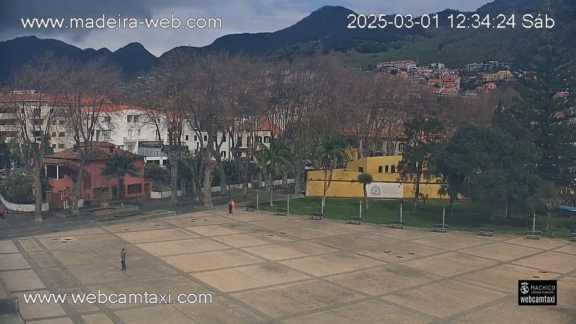 A paved plaza with a few people, surrounded by buildings and trees under a partly cloudy sky, with mountains in the background.