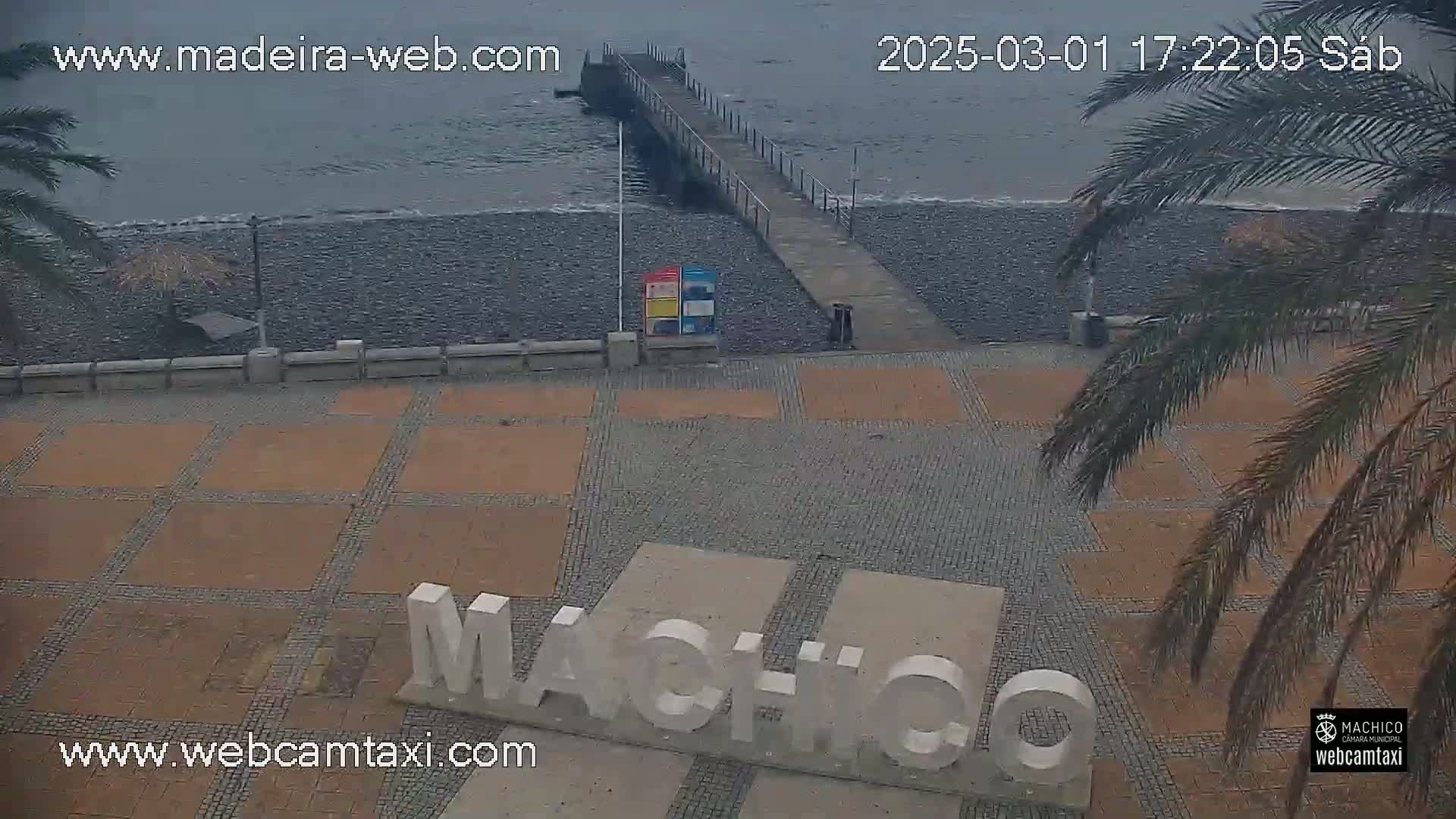 A paved plaza with a large "Machico" sign near a pebble beach and a pier, under an overcast sky.