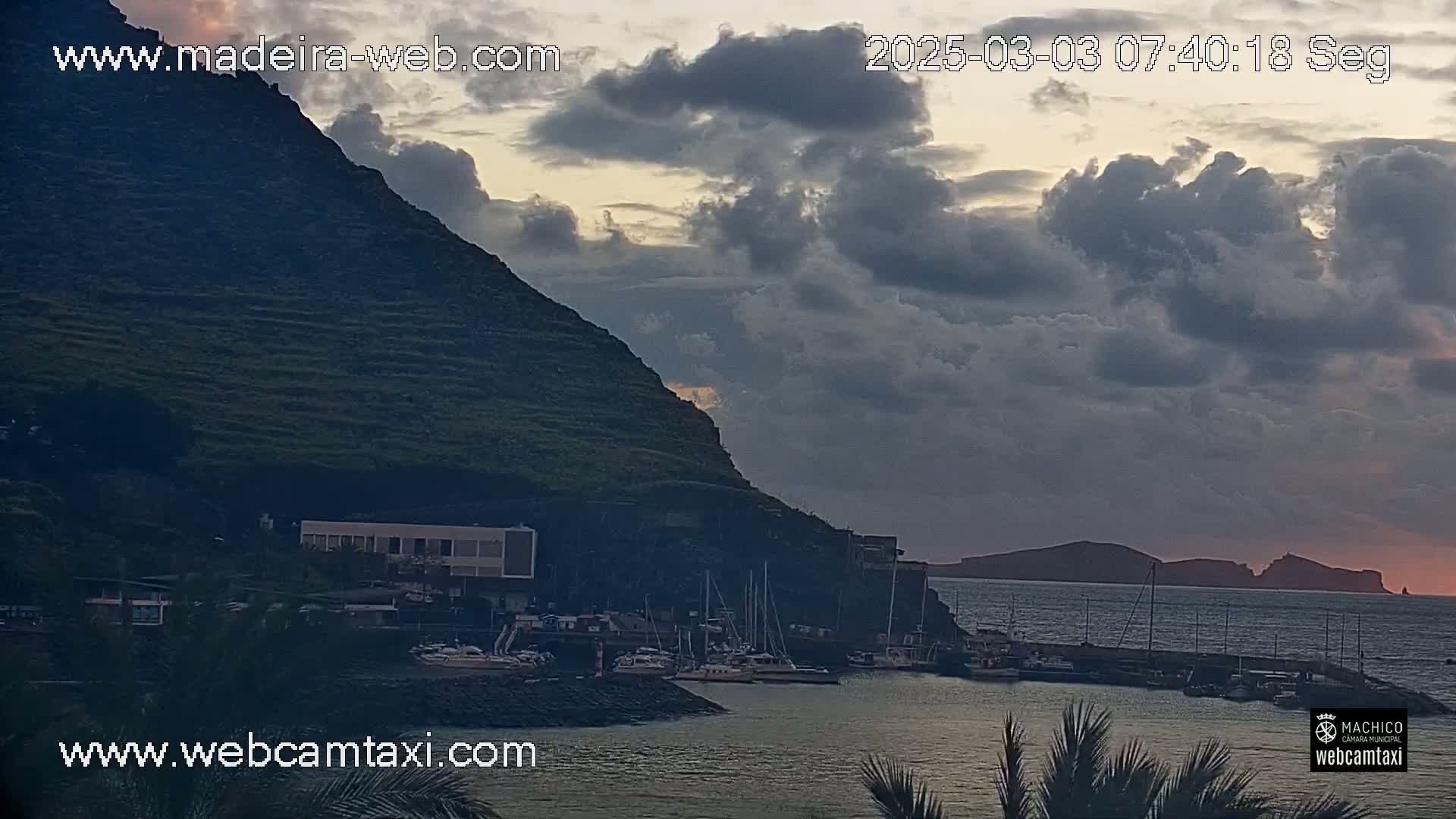 A harbor filled with boats sits below a dark, cloud-filled sky at sunset, nestled against a steep, terraced hillside.