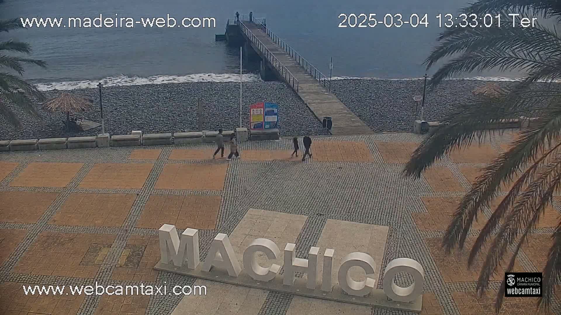 A paved plaza with large white letters spelling out a place name overlooks a pebble beach and a pier on an overcast day.