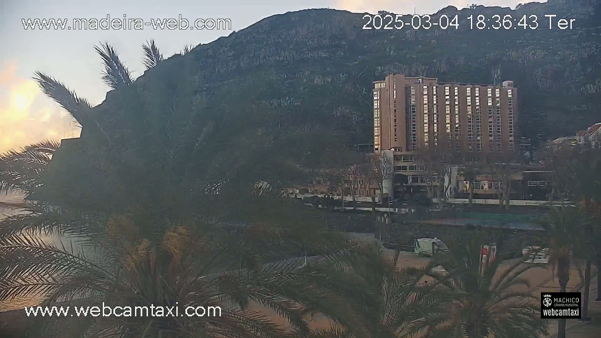 A large hotel is visible near a rocky cliff at sunset, with palm trees in the foreground.