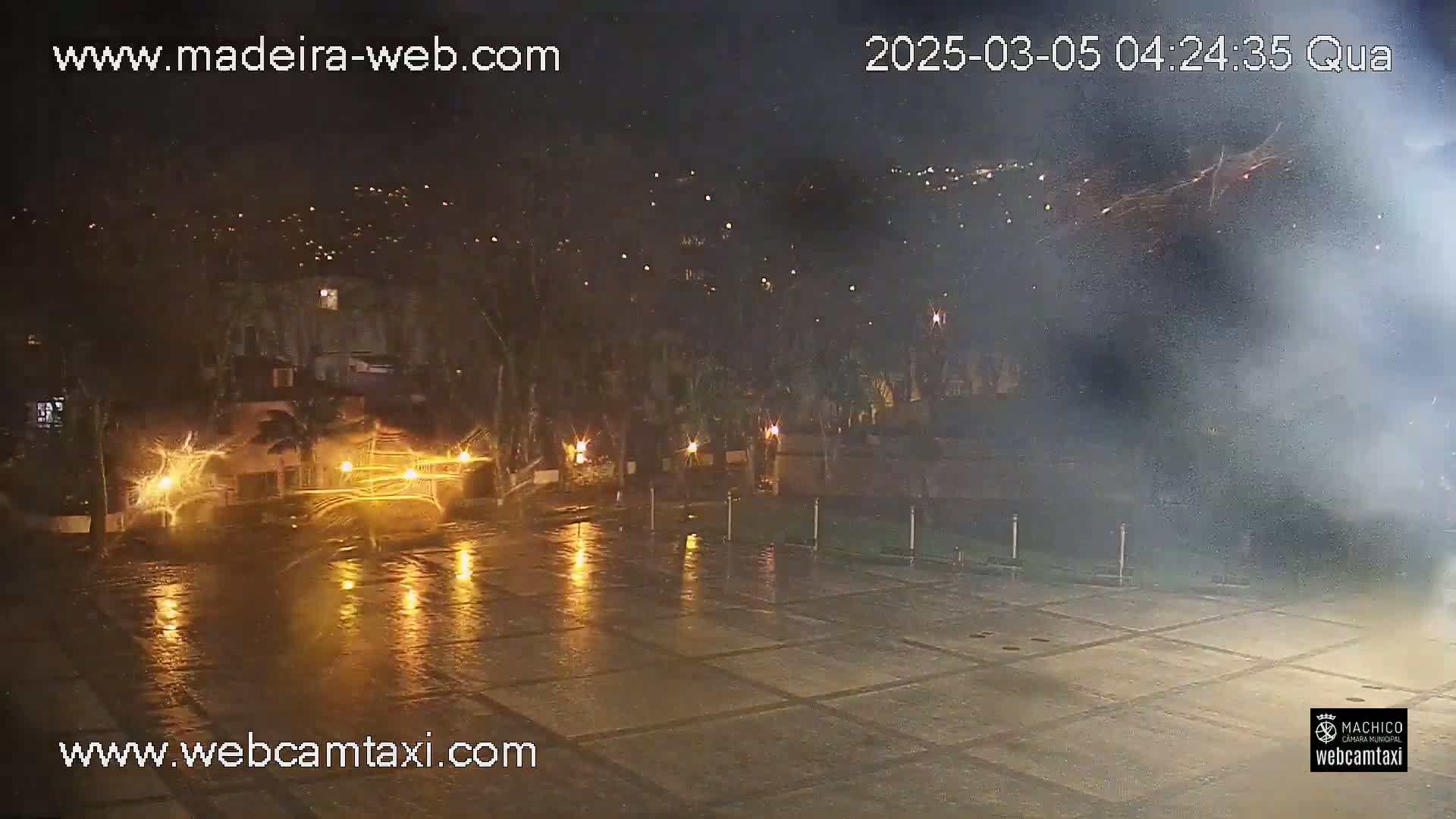 A rain-slicked nighttime plaza is illuminated by streetlights and distant lights reflecting on the wet pavement, with smoke or fog in the background.