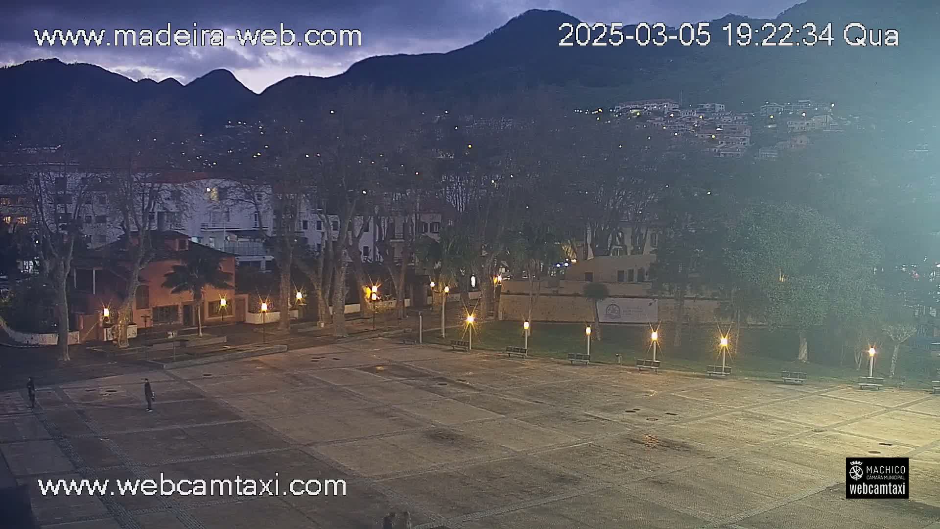 A nighttime view of a town square, illuminated by lampposts, with buildings and trees surrounding it, under a mostly cloudy, dark sky, and mountains in the background.
