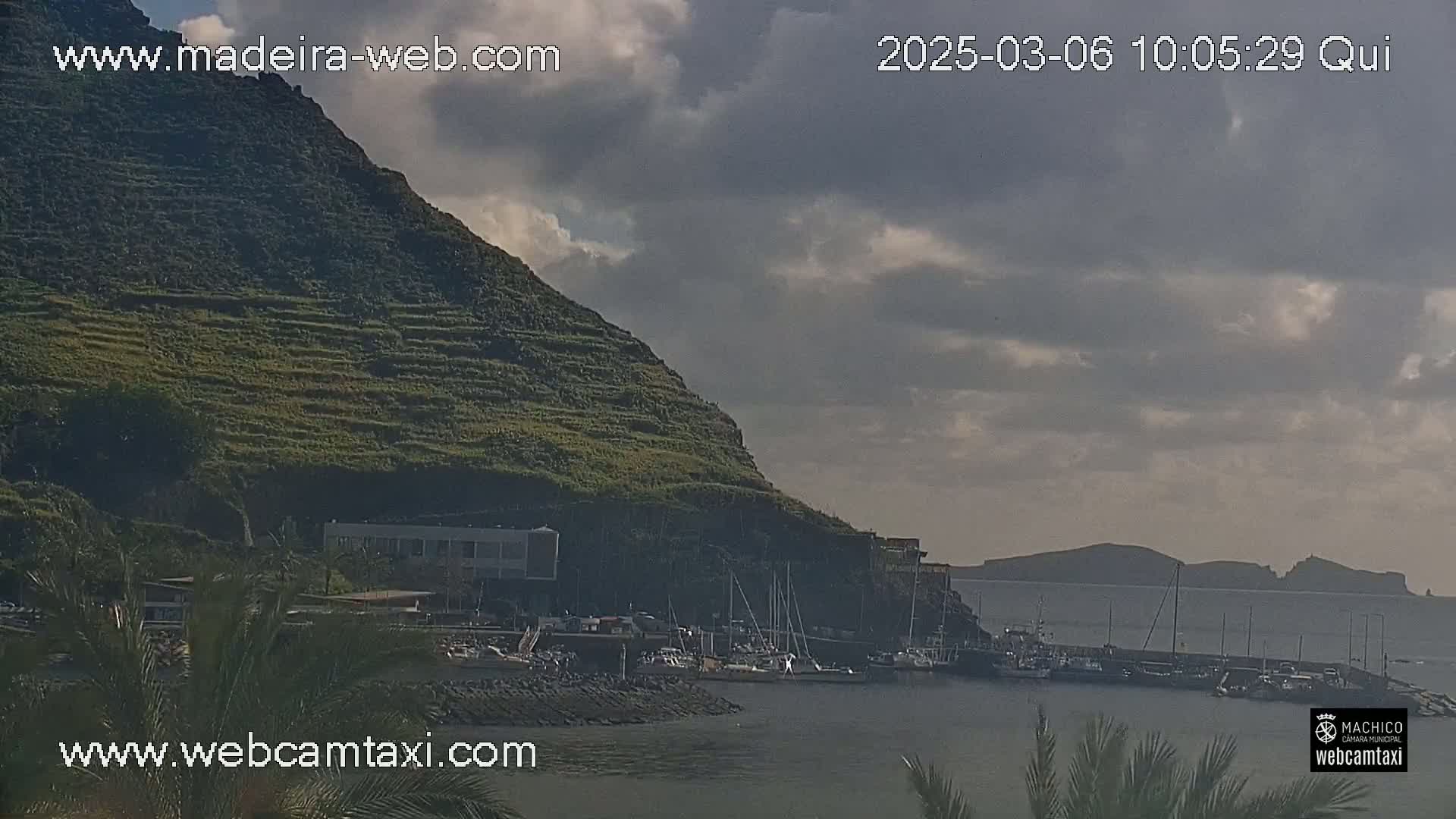 A partly cloudy day reveals a small harbor filled with boats nestled beneath a terraced hillside.