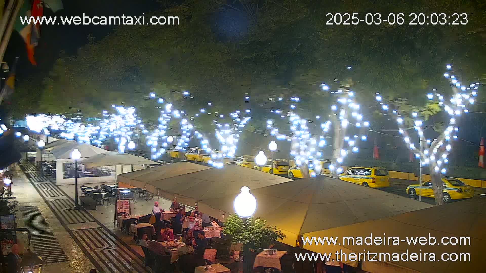At night, under string lights illuminating trees lining a street, yellow taxis are parked near outdoor cafe seating areas with patrons dining.