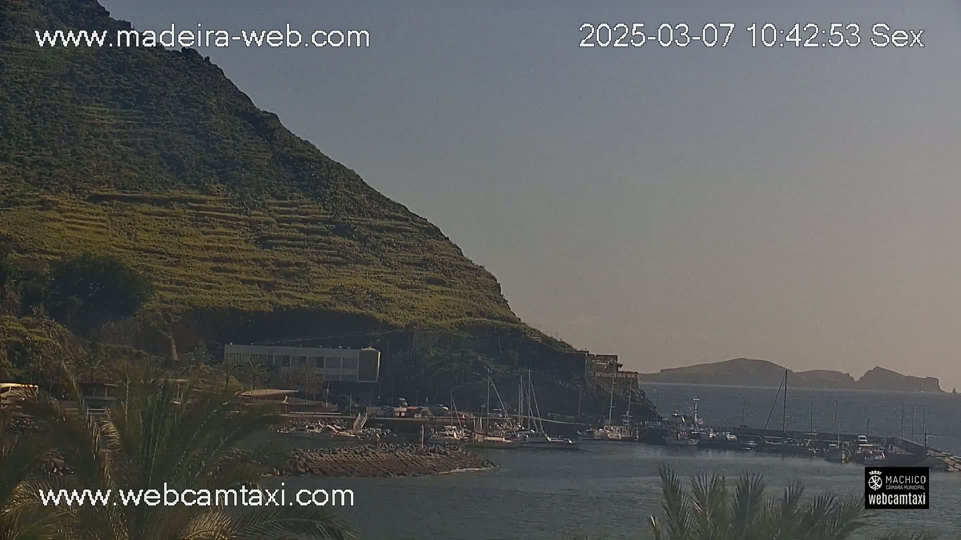 A hazy, sunny day reveals a harbor filled with boats nestled beside a steep, terraced hillside, with distant islands visible on the horizon.