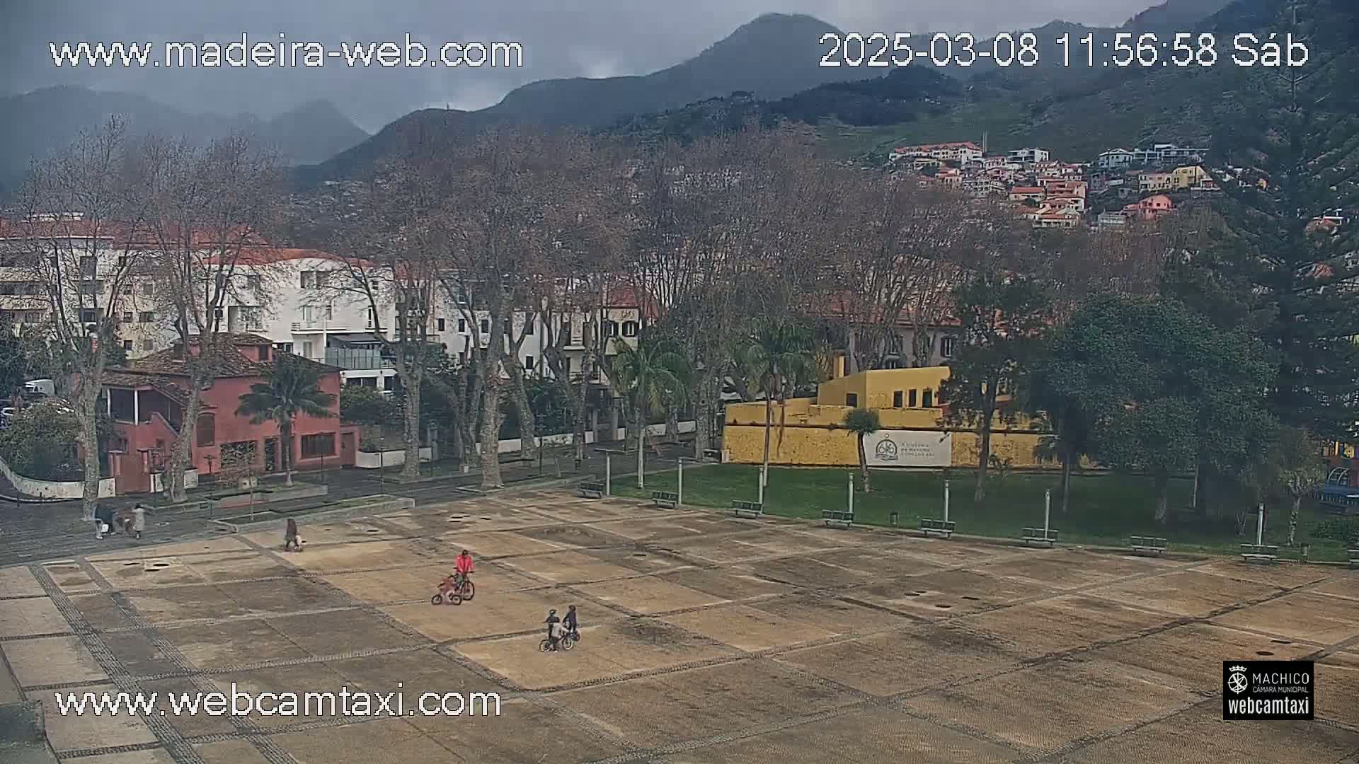A mostly overcast view of a town square with a few people and children on bicycles, surrounded by buildings and trees, with mountains in the background.