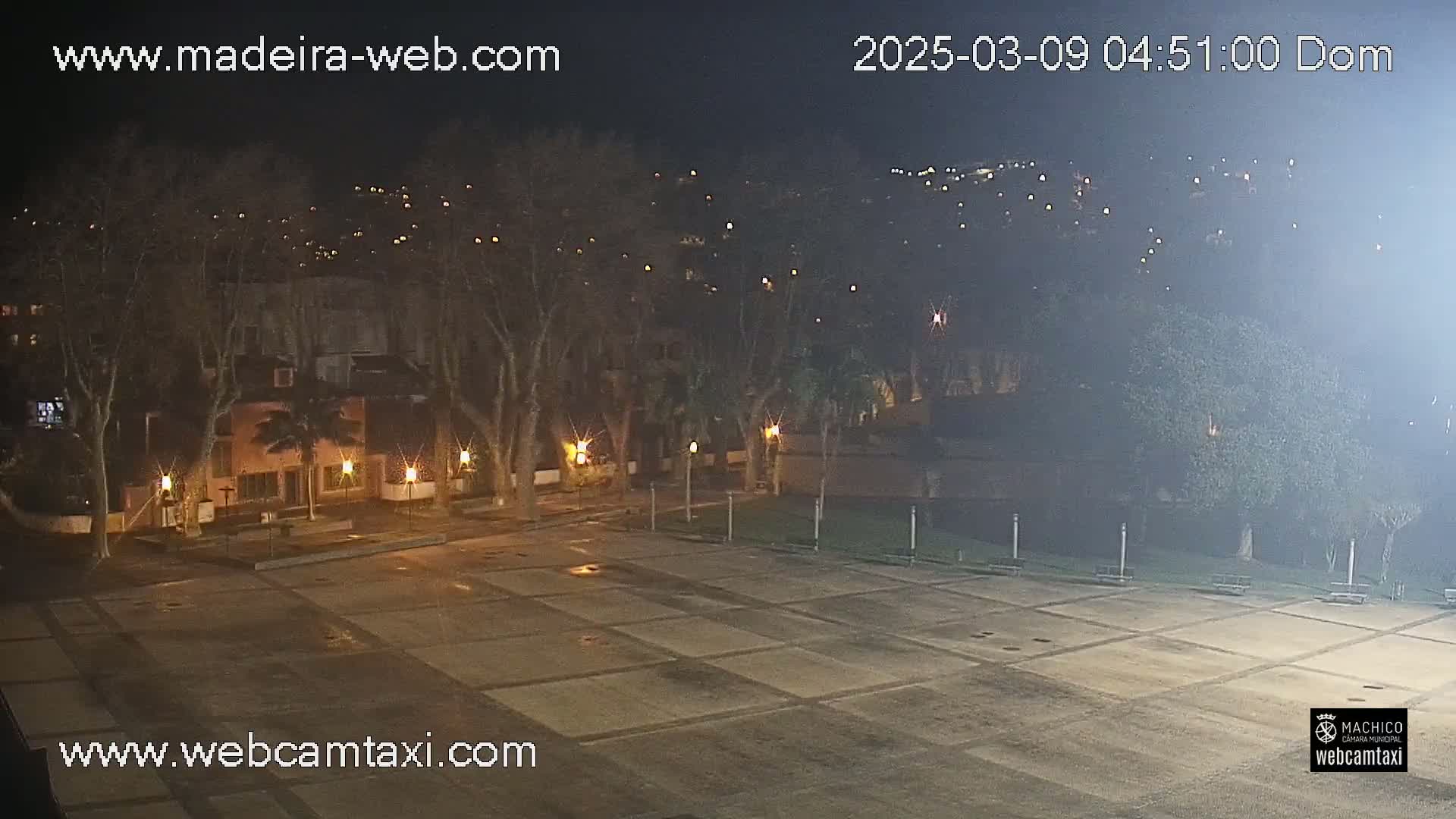 A nighttime view of a mostly empty plaza, surrounded by trees and buildings with lights, under a hazy sky.