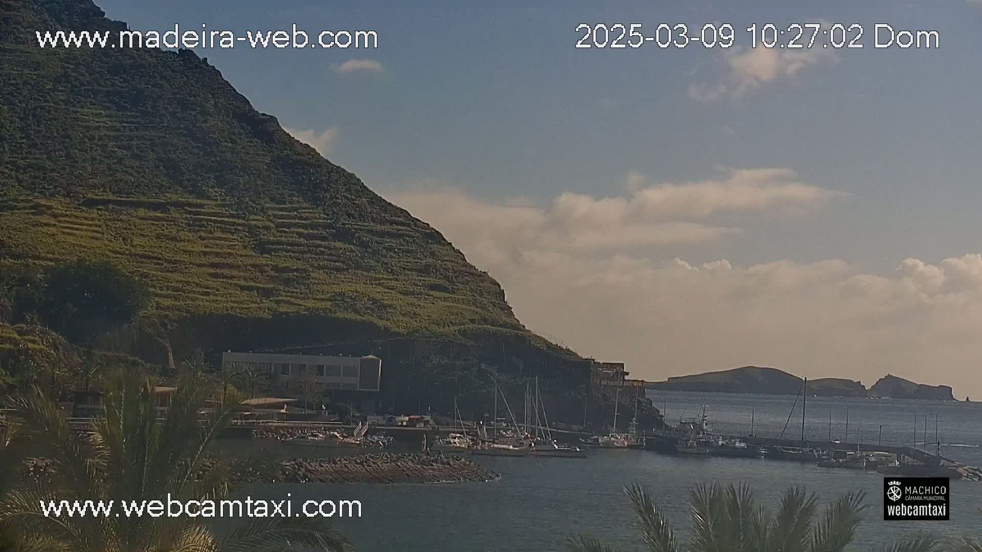 A partly cloudy day reveals a harbor filled with boats nestled beside a terraced hillside, with a small island visible in the distance.
