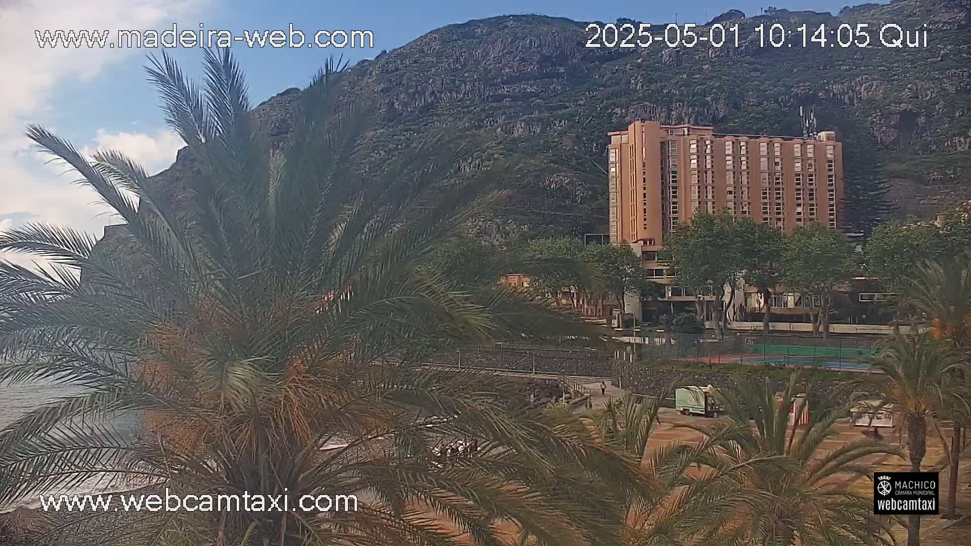 A partly cloudy day reveals a tall, light-brown building nestled amongst palm trees, near a rocky mountainside and a body of water.