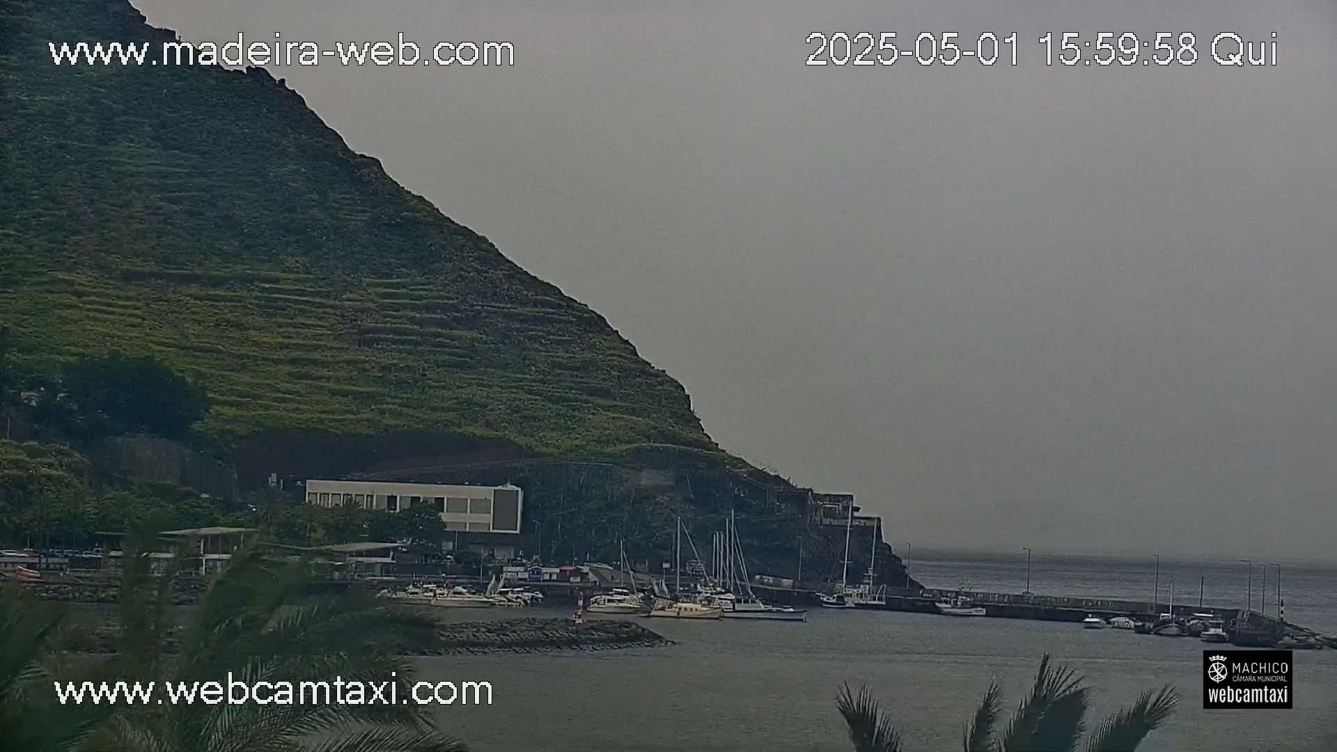 A hazy view shows a small harbor with several boats moored near a steep, terraced hillside.