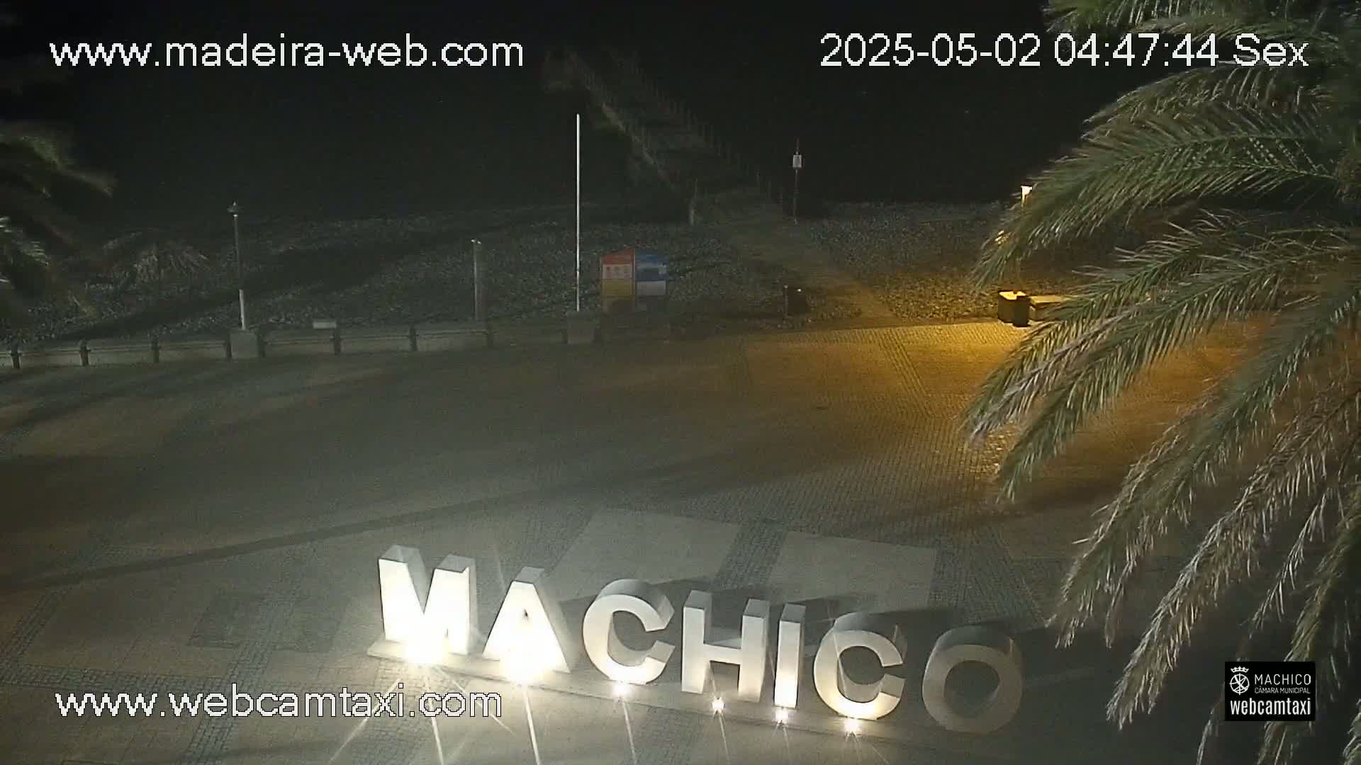 A nighttime, outdoor view of a plaza with the word "MACHICO" displayed in large letters, palm trees, and a dark beach in the background.