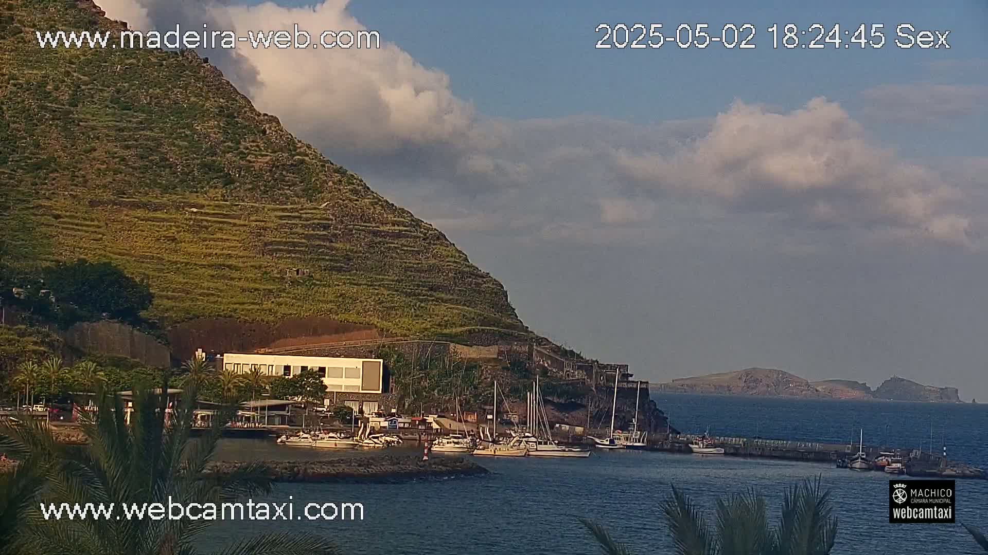 A partly cloudy day reveals a marina nestled beside a terraced hillside, with a small island visible in the distance.