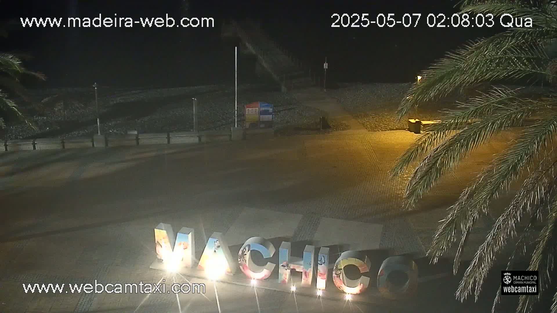 A nighttime view of a beach area with a large "Machico" sign illuminated, palm trees, and a dark, pebble-covered shoreline.